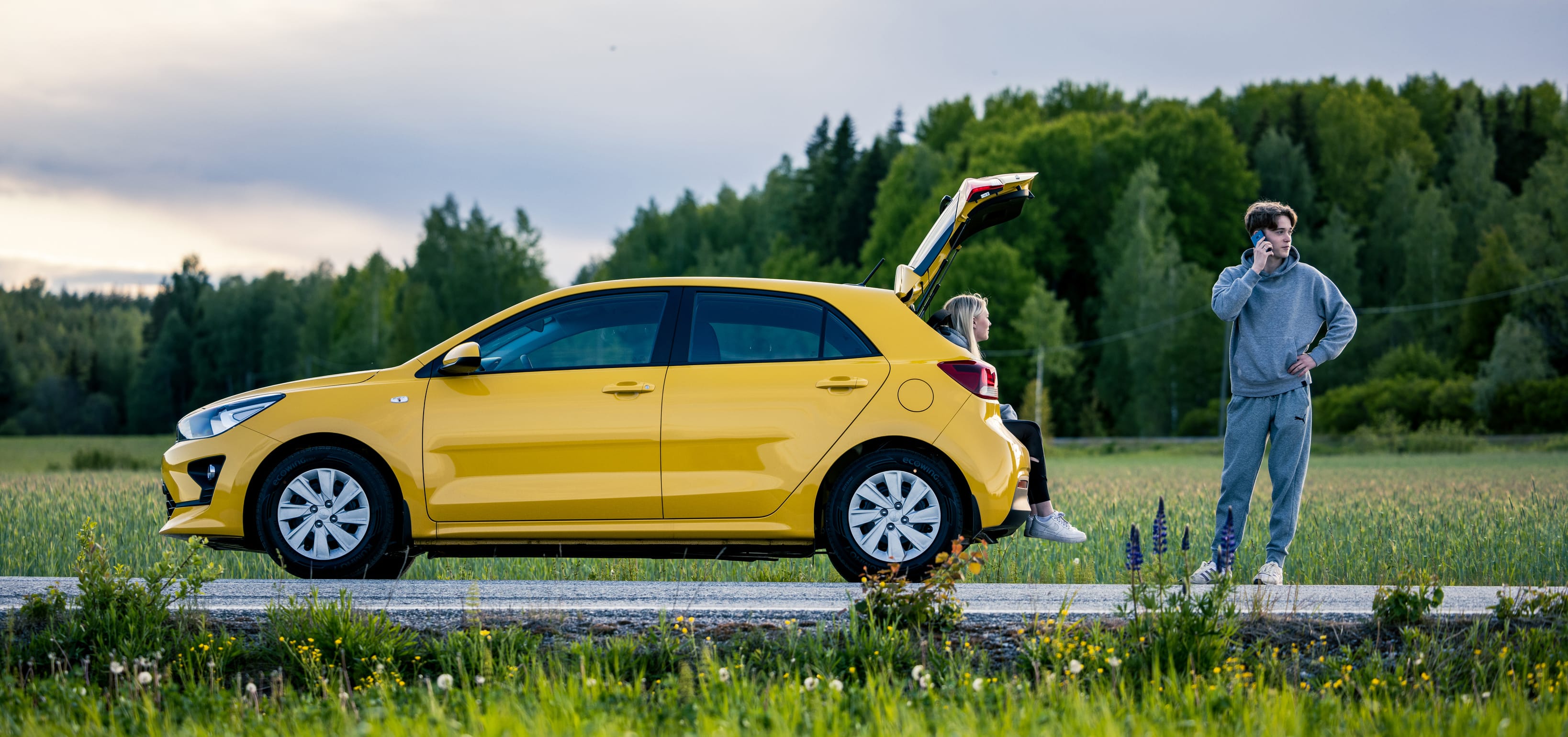 Keltainen auto, takaluukku auki. Tyttö istuu takaluukussa, poika tiellä puhelimessa. Autossa ongelma.